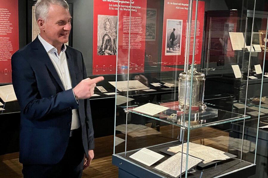 Ein Mann im Anzug zeigt in einem Museum auf eine Glasvitrine mit Exponaten.