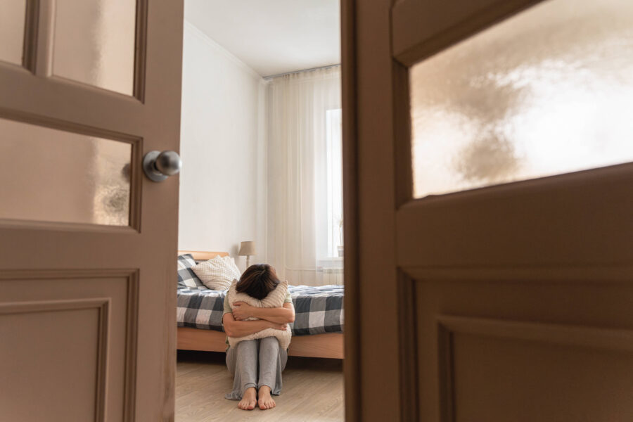 Offene Tür, Person sitzt mit Kissen auf dem Boden neben einem Bett.