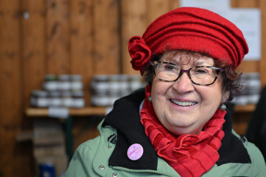 Ältere Frau mit roter Mütze und Brille lächelt, trägt Anstecker.