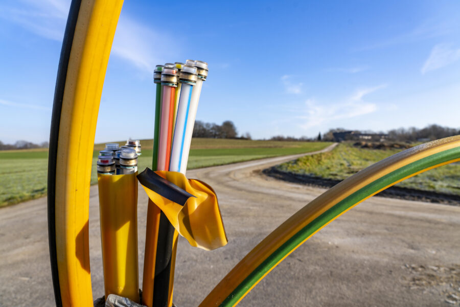 Bunte Glasfaserkabel ragen aus gelbem Schutzrohr vor ländlichem Weg.