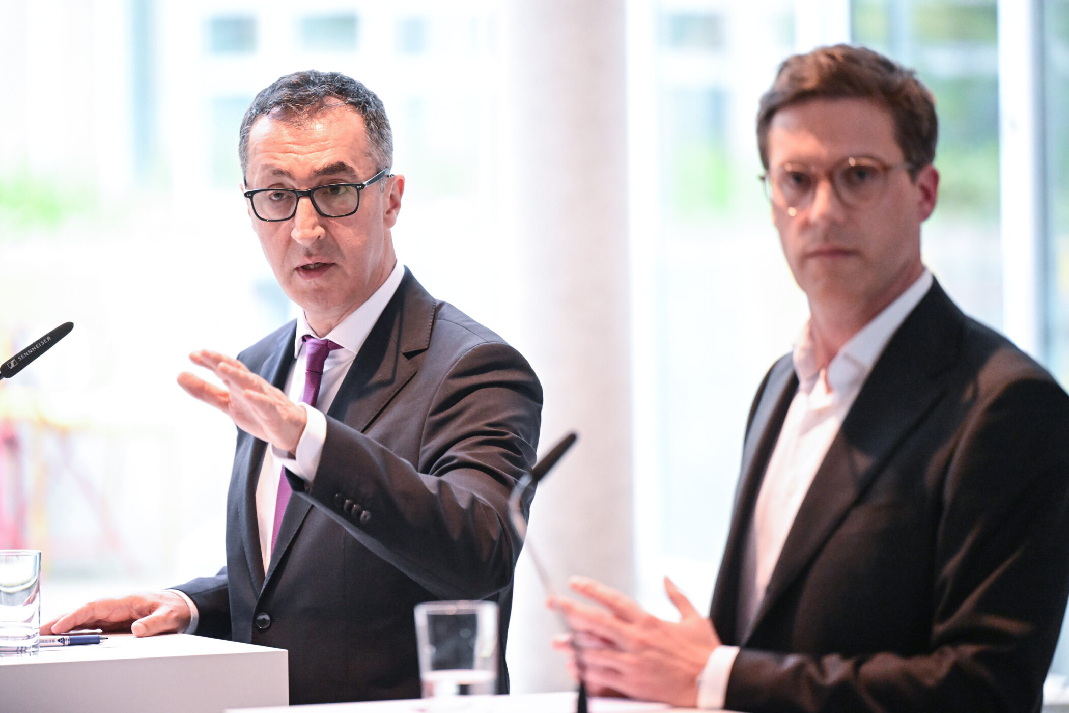 Zwei Männer im Anzug sprechen an Mikrofonen auf einem Podium.