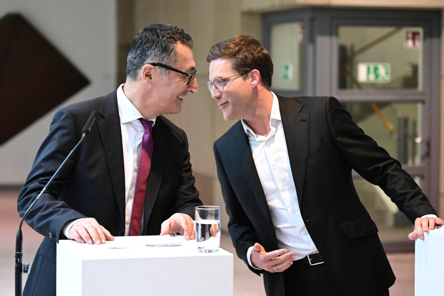 Zwei Männer in Anzügen stehen lächelnd an weißen Pulten mit Mikrofon.