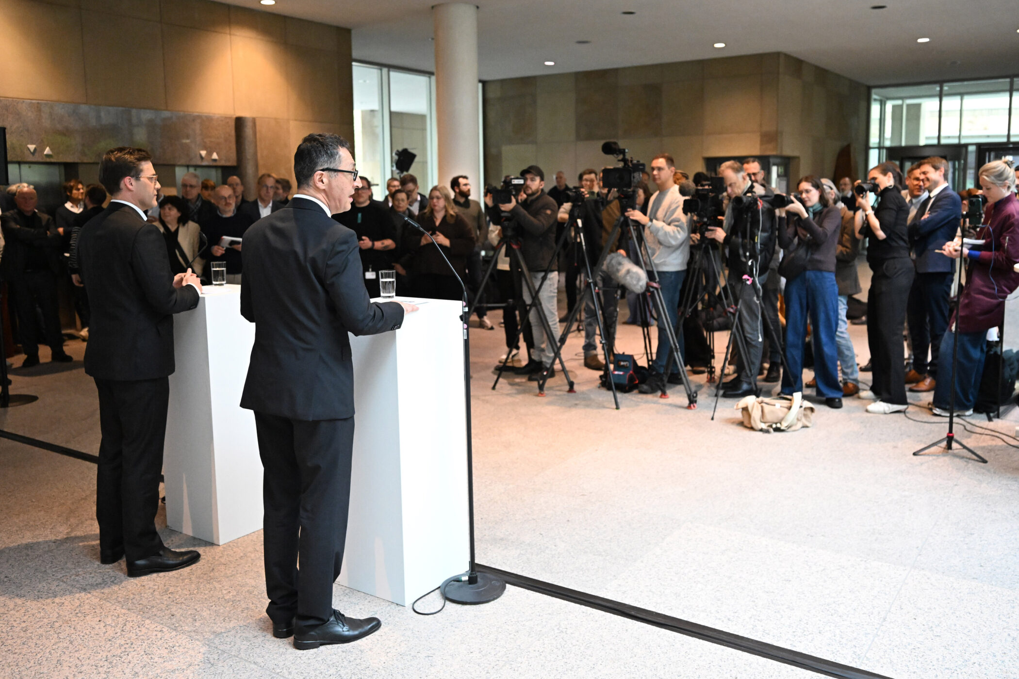 Zwei Männer stehen an weißen Podesten, umgeben von Kameraleuten und Publikum.