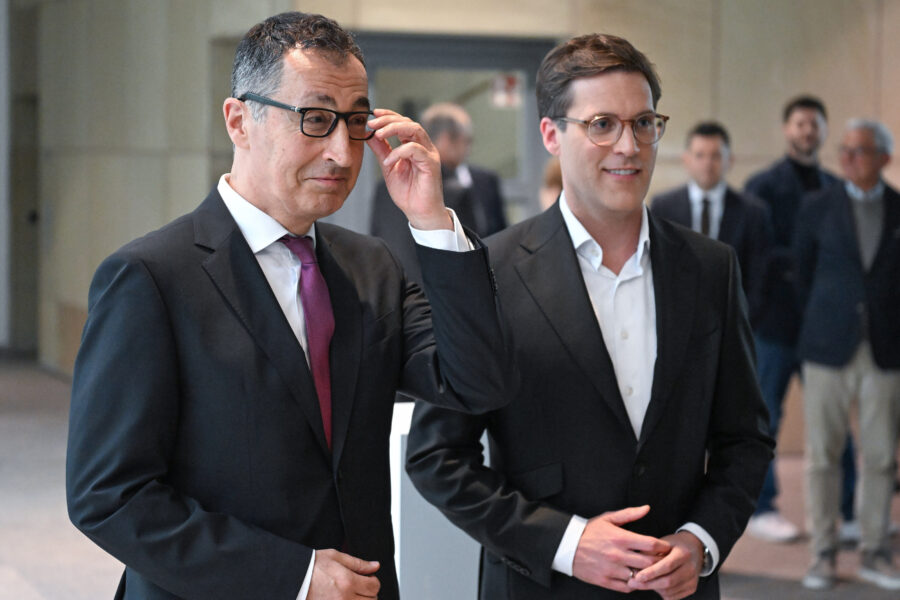 Zwei Männer in Anzügen stehen nebeneinander, Hintergrund mit mehreren Personen.