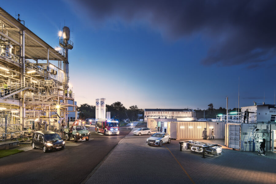 Industrieanlage bei Dämmerung, beleuchtet, verschiedene Fahrzeuge, Personen in Bewegung.