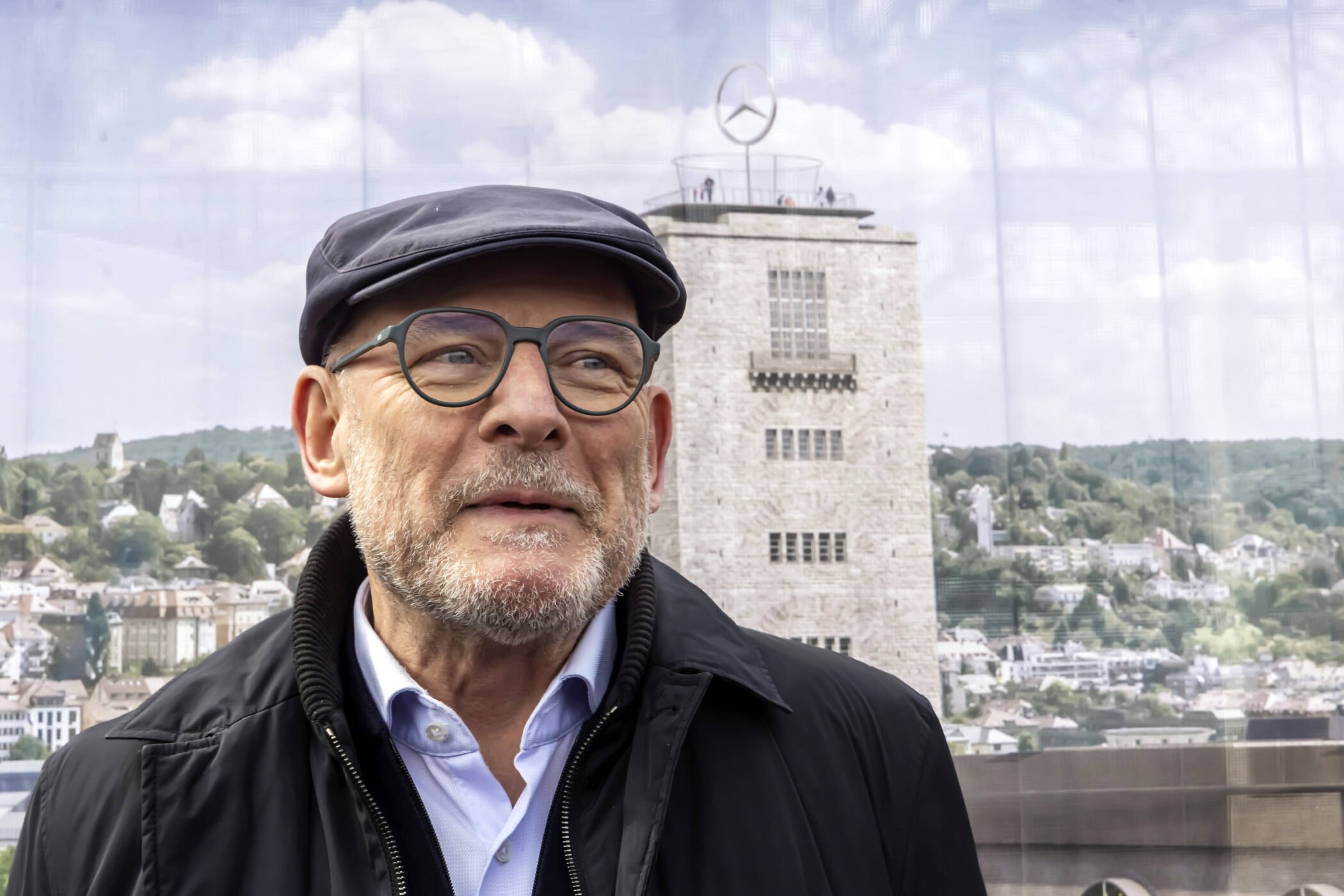 Mann mit Brille und Mütze vor einem Turm mit Mercedes-Logo, Stadtansicht im Hintergrund.