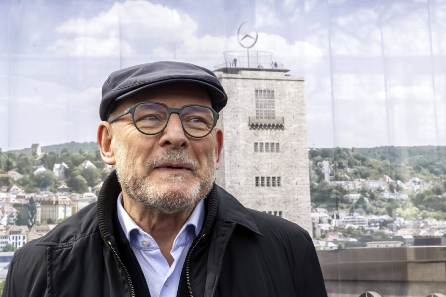 Mann mit Brille und Mütze vor einem Turm mit Mercedes-Logo, Stadtansicht im Hintergrund.