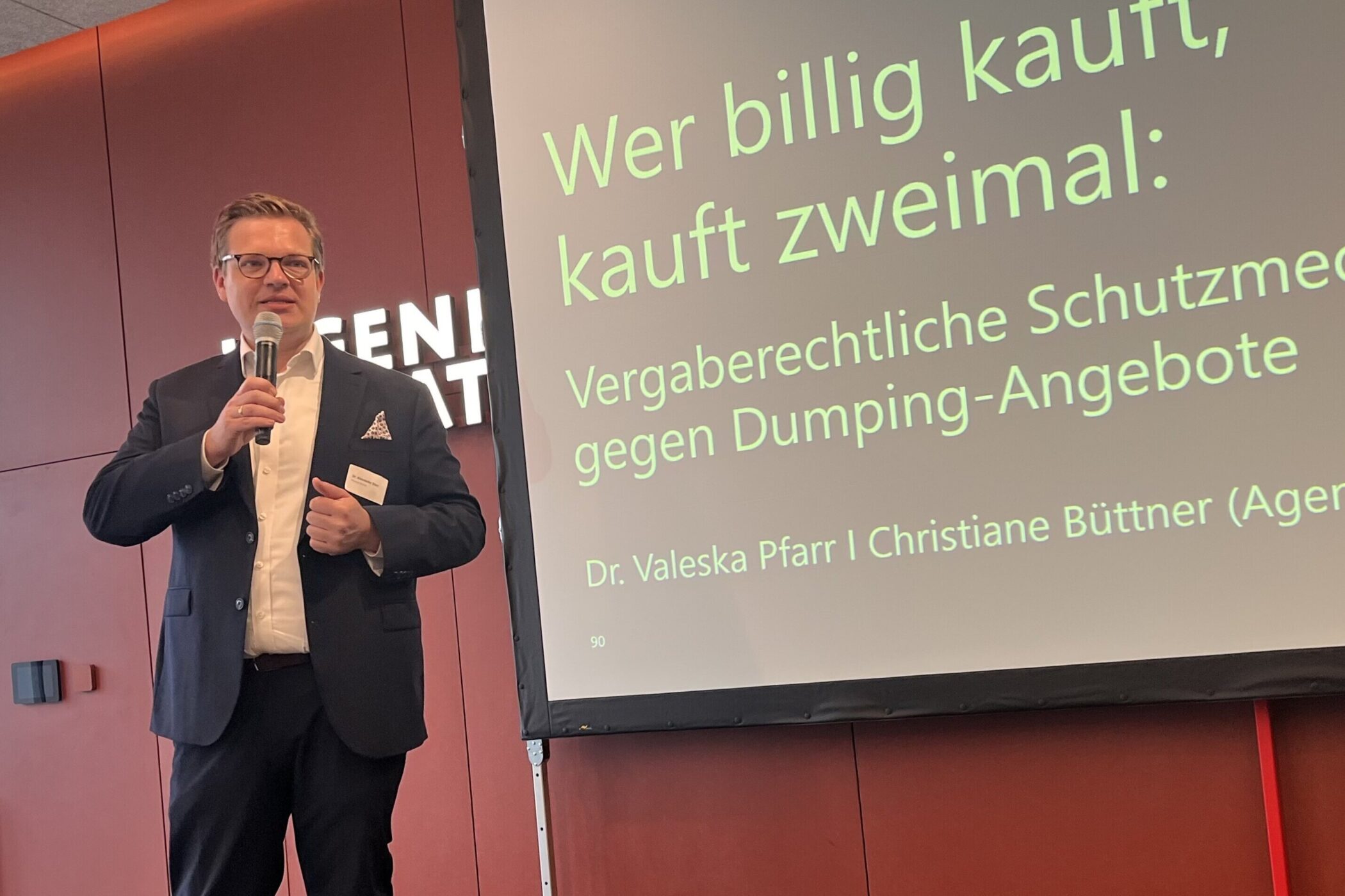 Mann hält Mikrofon, Leinwand zeigt Text über Vergabeschutz gegen Dumping-Angebote.