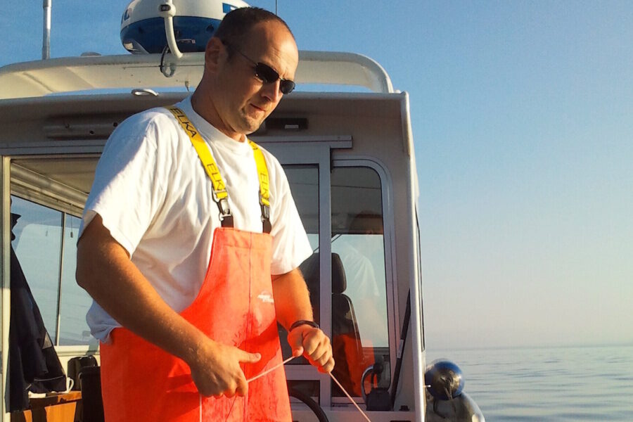 Mann mit orangefarbener Latzhose und Sonnenbrille auf einem Boot.