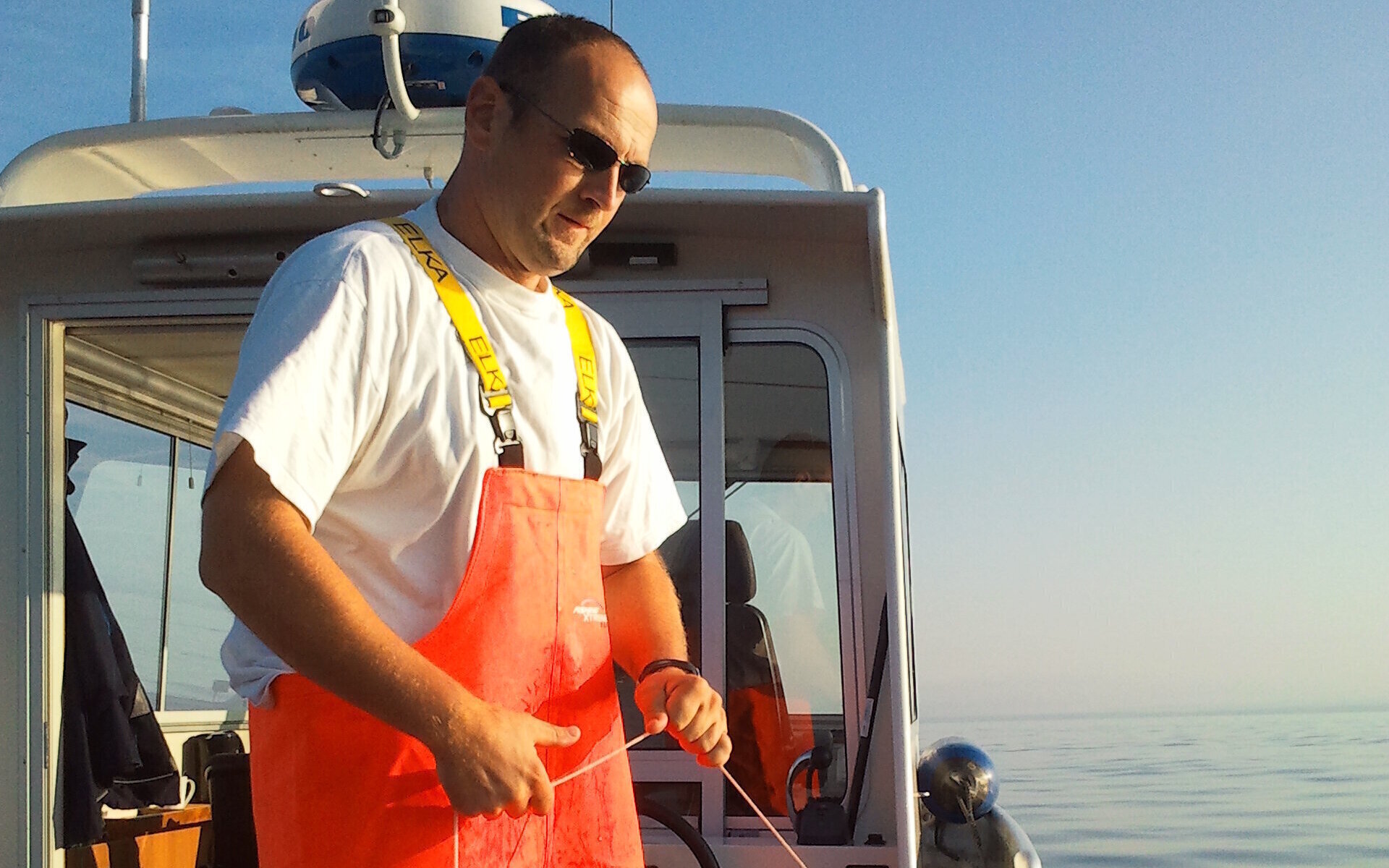 Mann mit orangefarbener Latzhose und Sonnenbrille auf einem Boot.