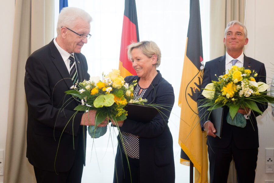 Drei Personen mit Blumensträußen vor deutschen Flaggen.