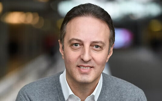 Ein lächelnder Mann in einem grauen Pullover und weißem Hemd.