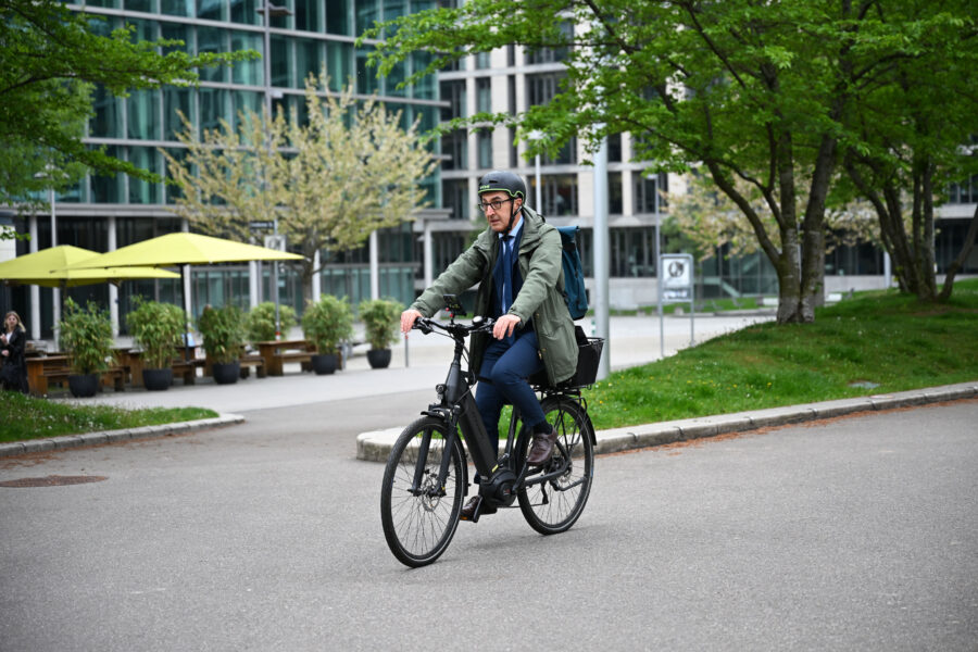 Mann mit Rucksack fährt Fahrrad in urbaner Umgebung, Bäume im Hintergrund.