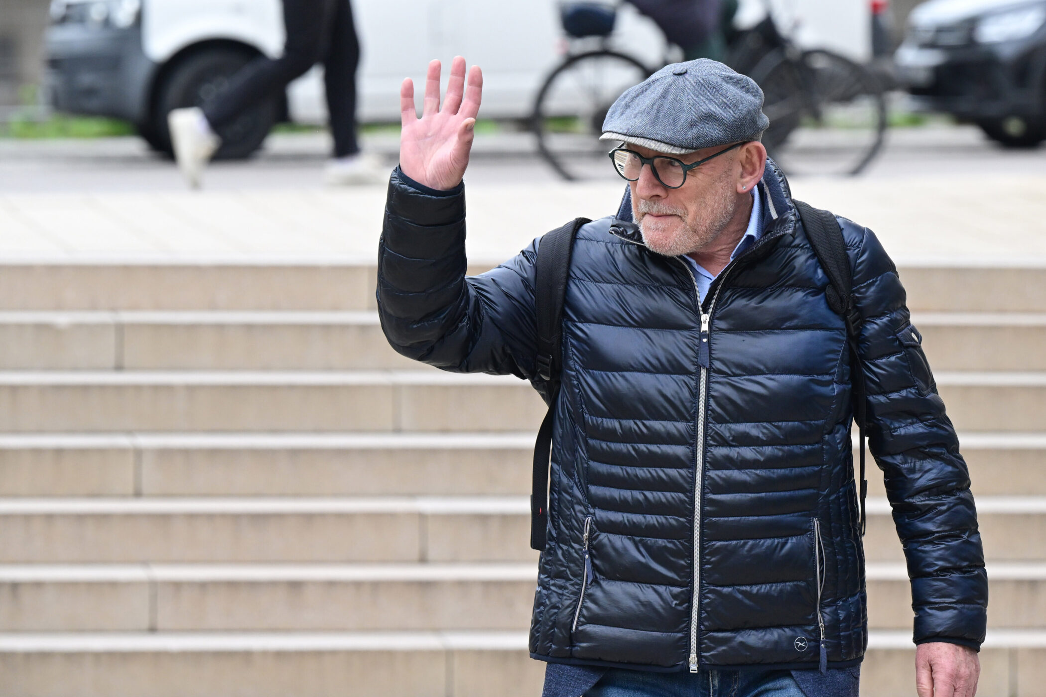 Mann in blauer Jacke hebt Hand, Treppe und Radfahrer im Hintergrund.