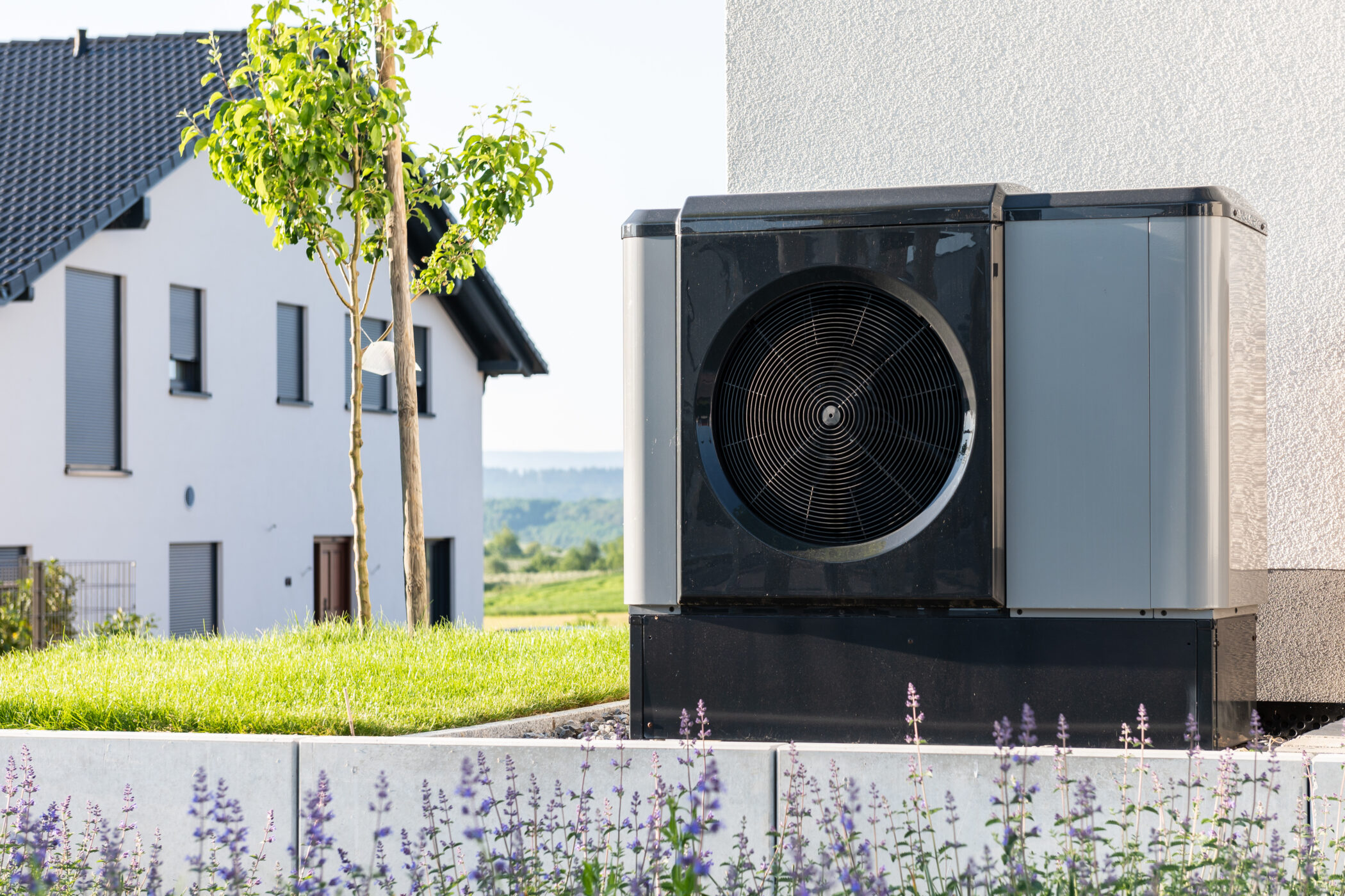 Luftwärmepumpe neben einem modernen Haus, im Vordergrund blühende Pflanzen.