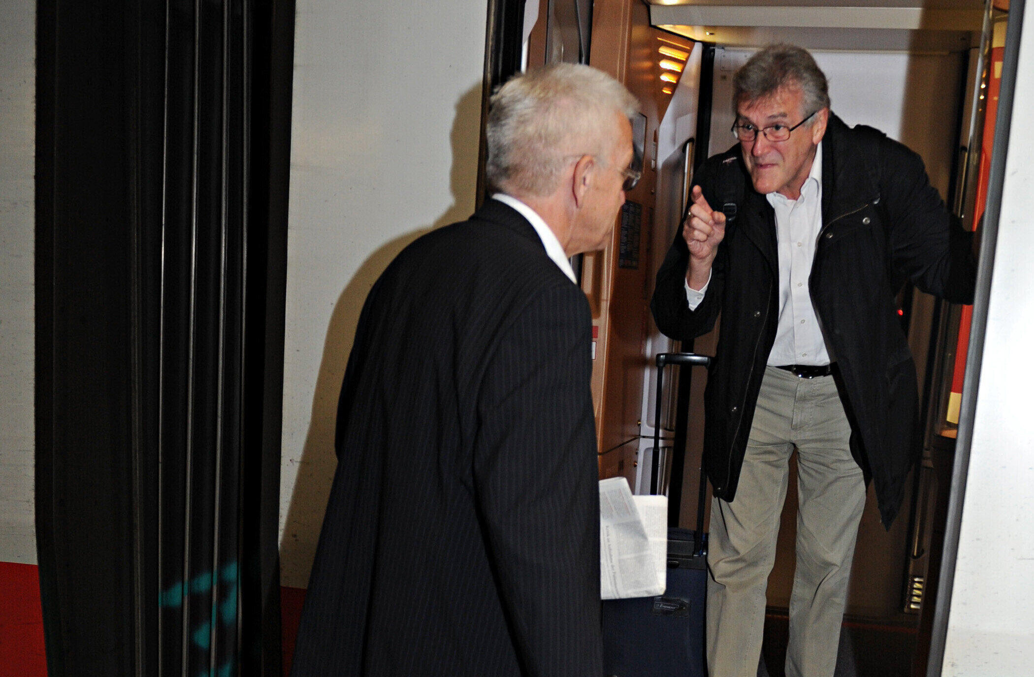 Zwei Männer sprechen an einem Zug, einer hält Zeitung und Koffer.