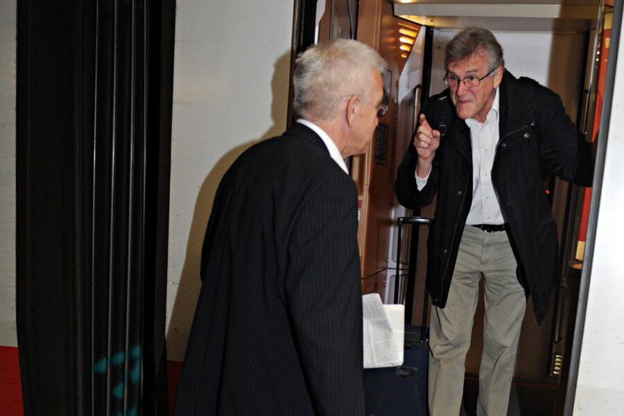 Zwei Männer sprechen an einem Zug, einer hält Zeitung und Koffer.