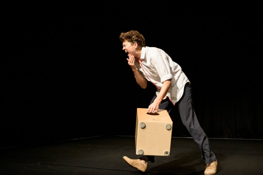 Person auf der Bühne mit weißem Hemd hält einen Holzwürfel und schreit.