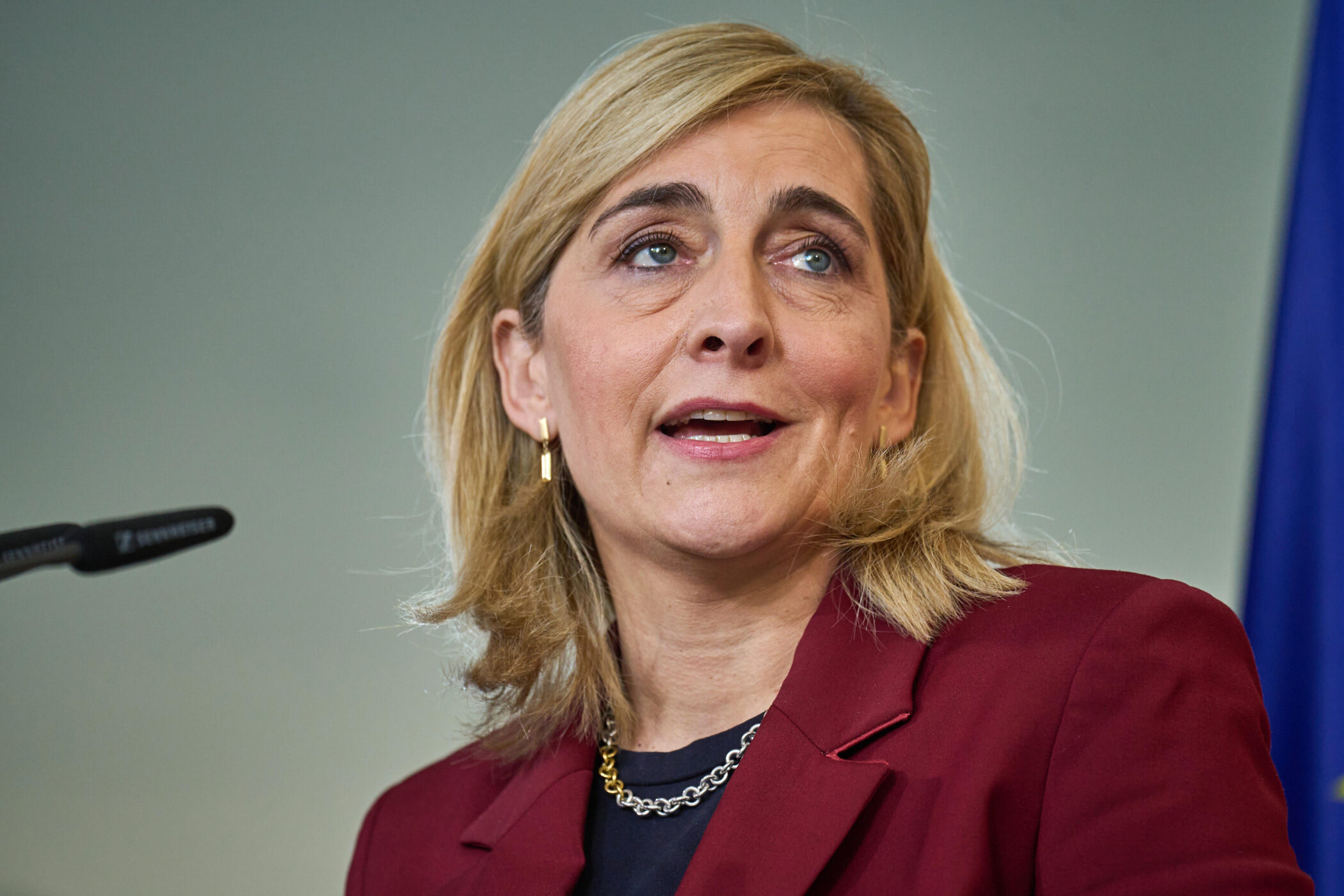 Frau in rotem Blazer steht vor Mikrofon, blaue Flagge im Hintergrund.