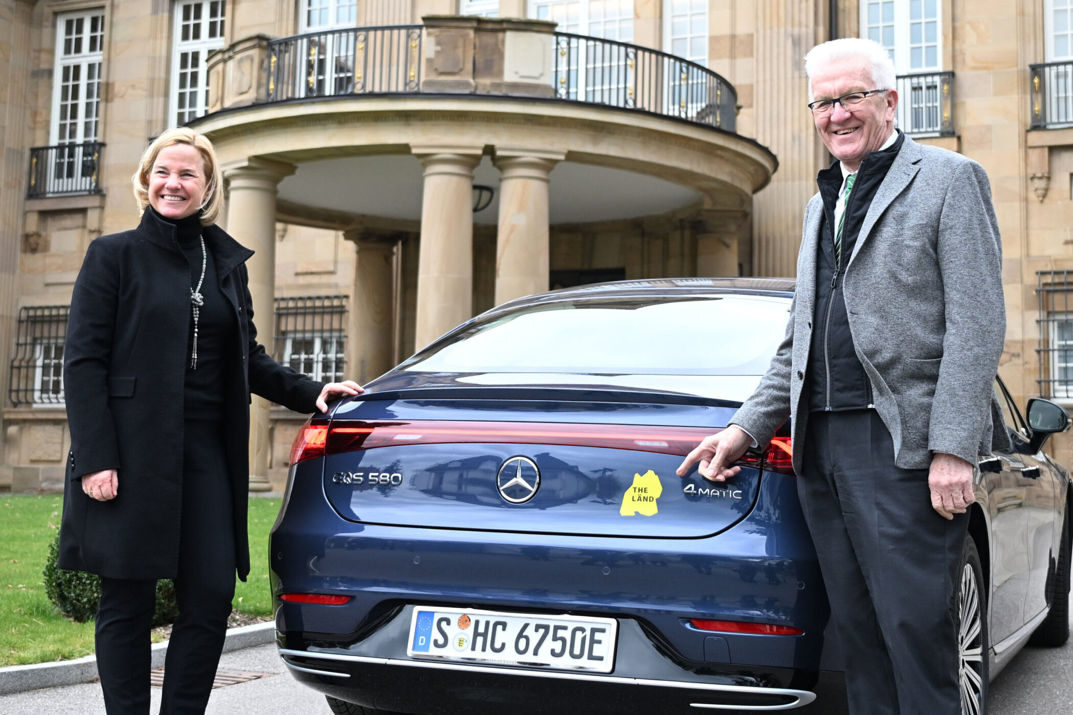 Zwei Personen vor einem Auto mit "THE LÄND" Aufkleber, Nummernschild S-HC 6750E.