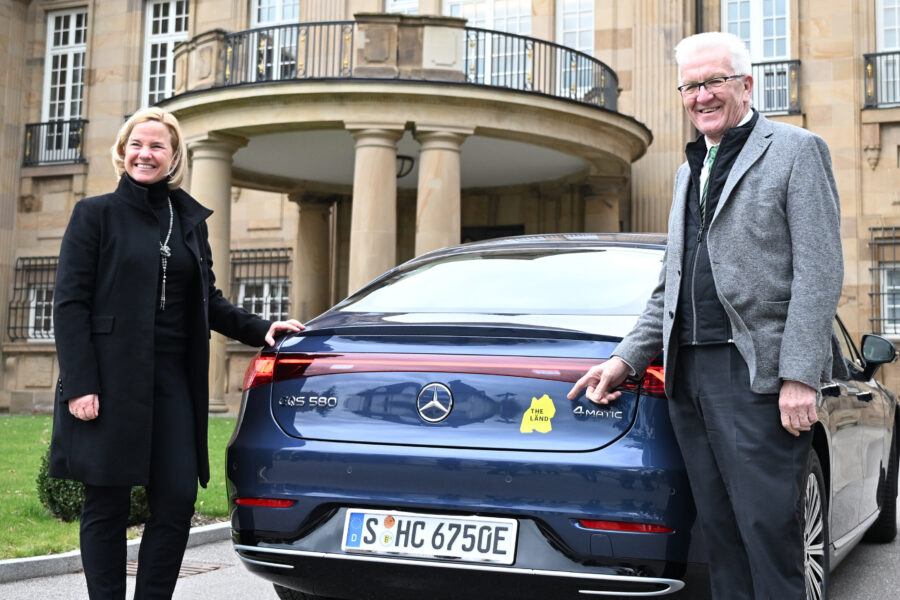 Zwei Personen vor einem Auto mit "THE LÄND" Aufkleber, Nummernschild S-HC 6750E.
