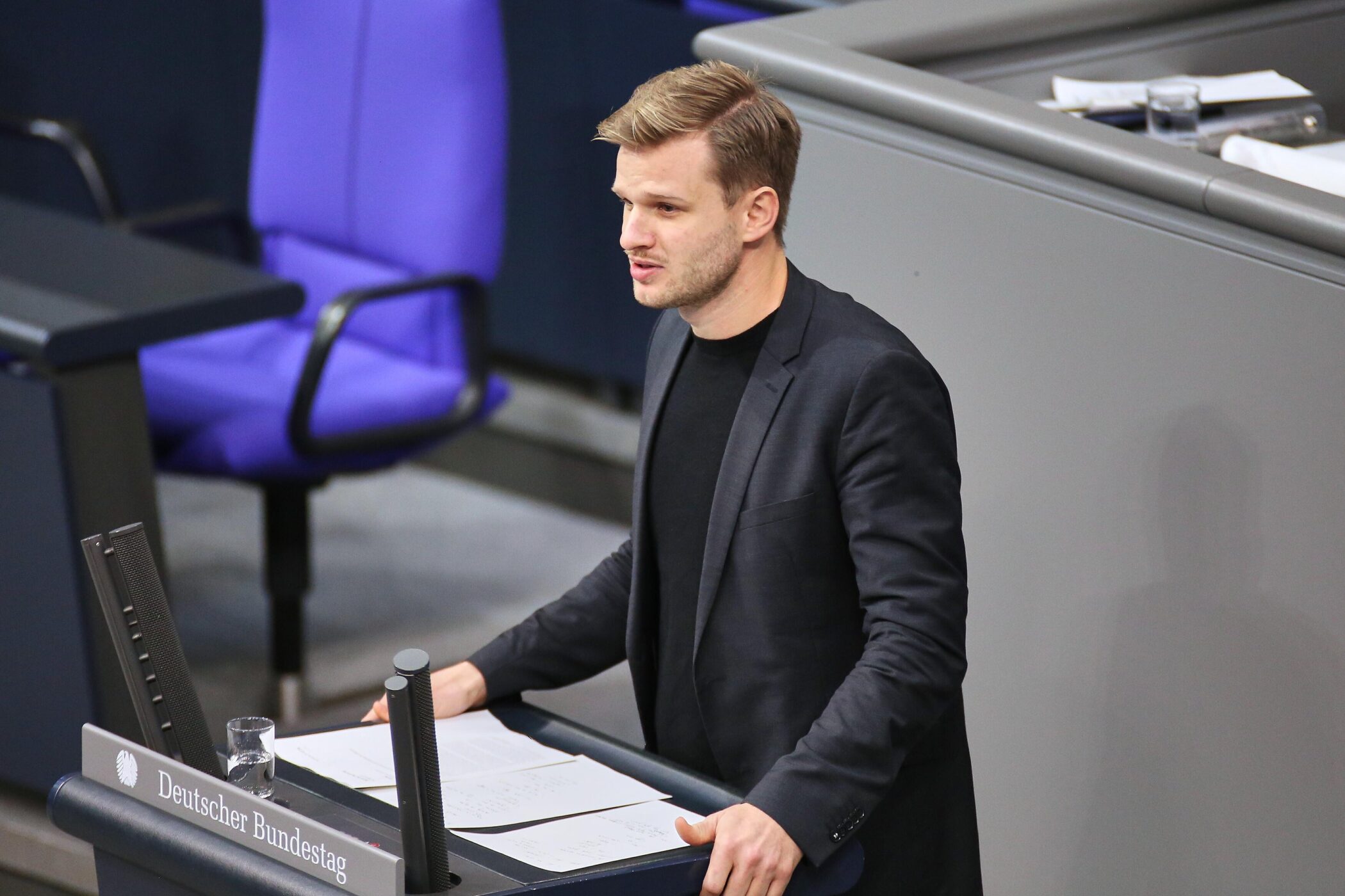 Mann am Rednerpult im Bundestag, blauer Stuhl im Hintergrund.