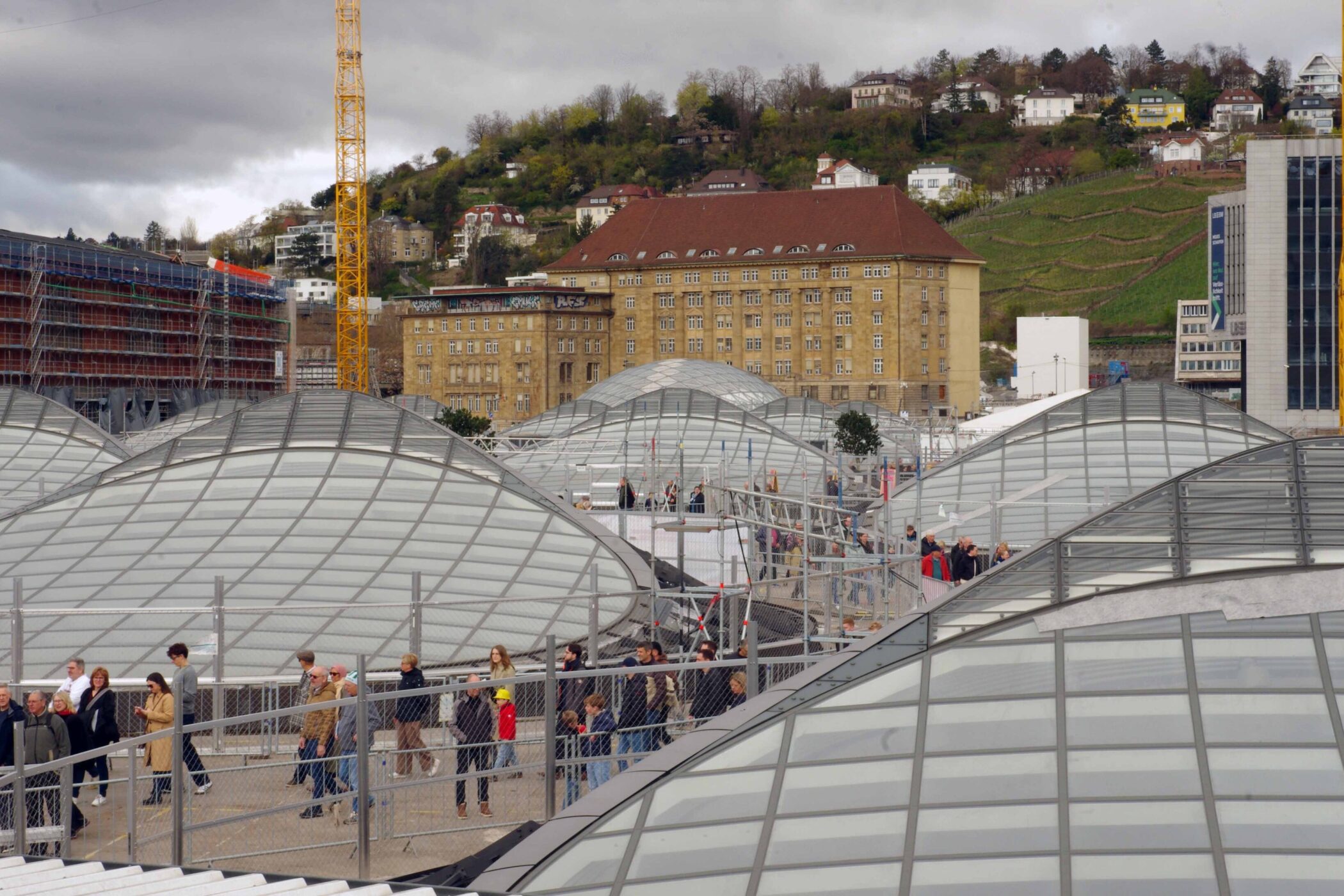 Personengruppe auf begehbarer Glasdachkonstruktion, Gebäude und Hügel im Hintergrund.