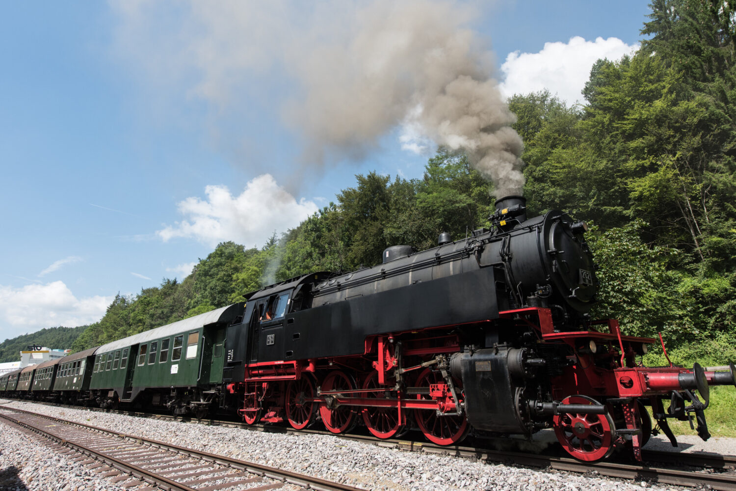 Dampflokomotive zieht Personenwagen durch eine grüne Waldlandschaft.