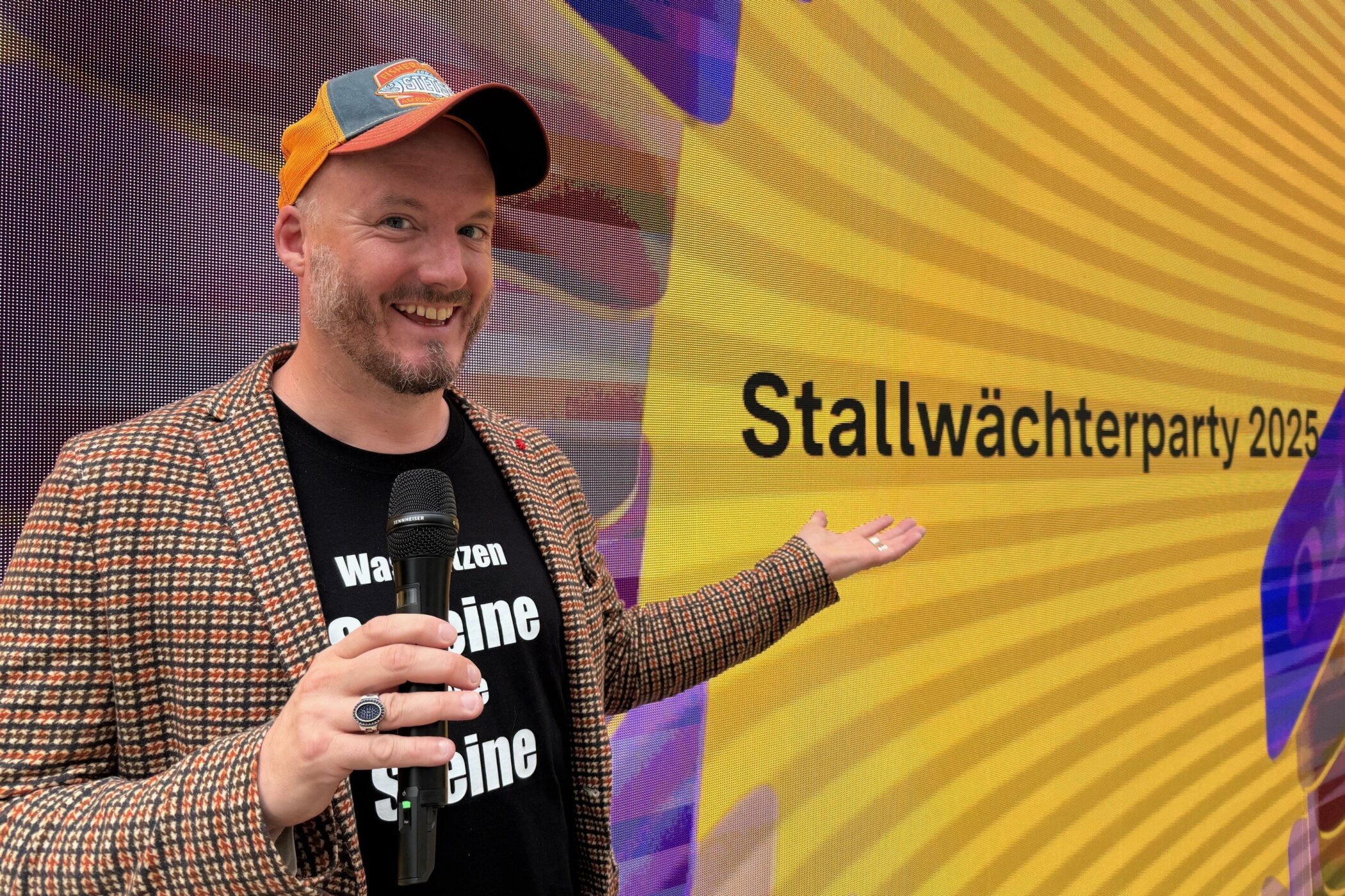 Mann mit Mikrofon vor gelbem Hintergrund mit Text: "Stallwächterparty 2025".