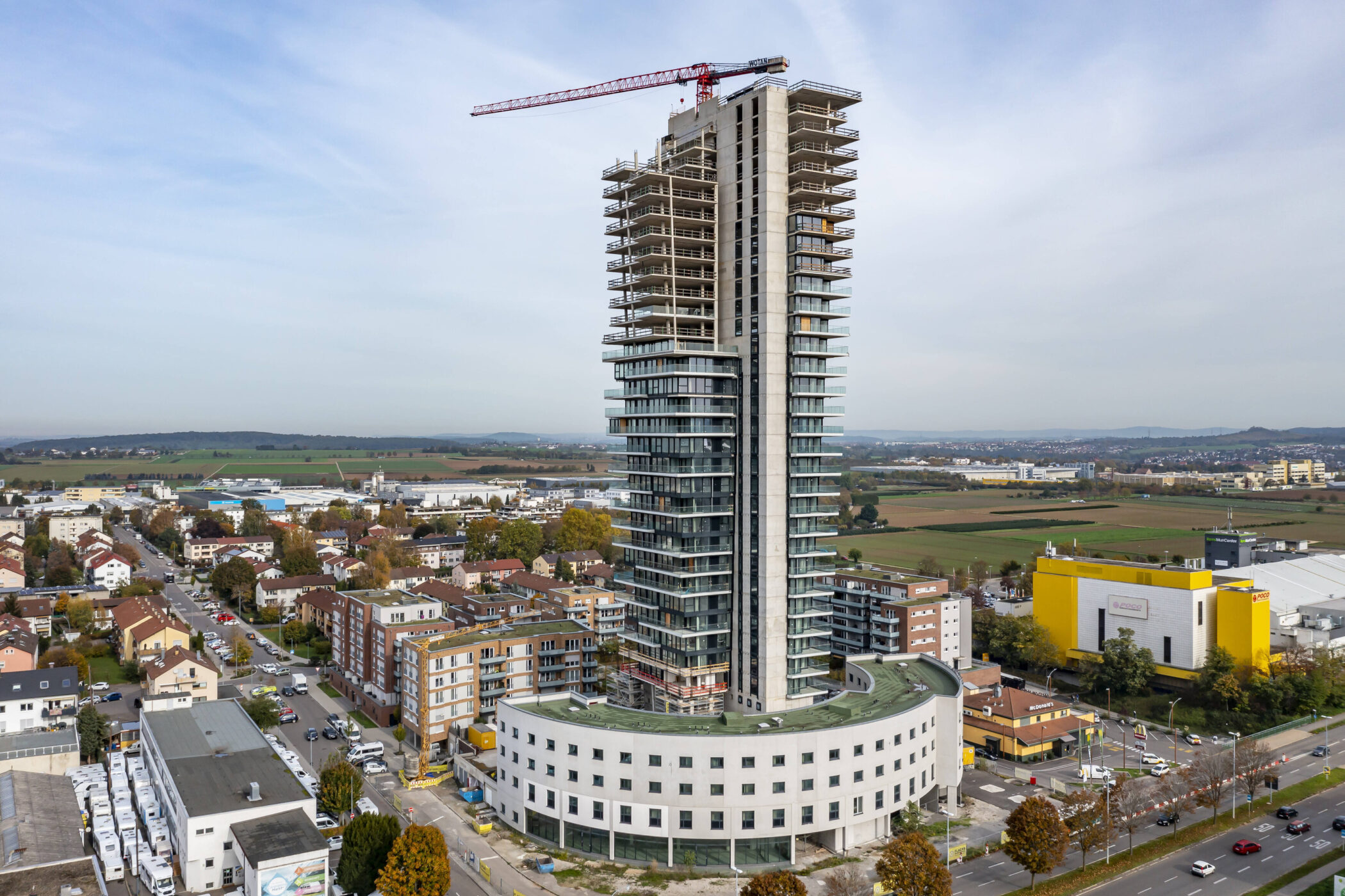 Hochhaus mit Kran in Stadtumgebung, umliegende Gebäude und Felder im Hintergrund.