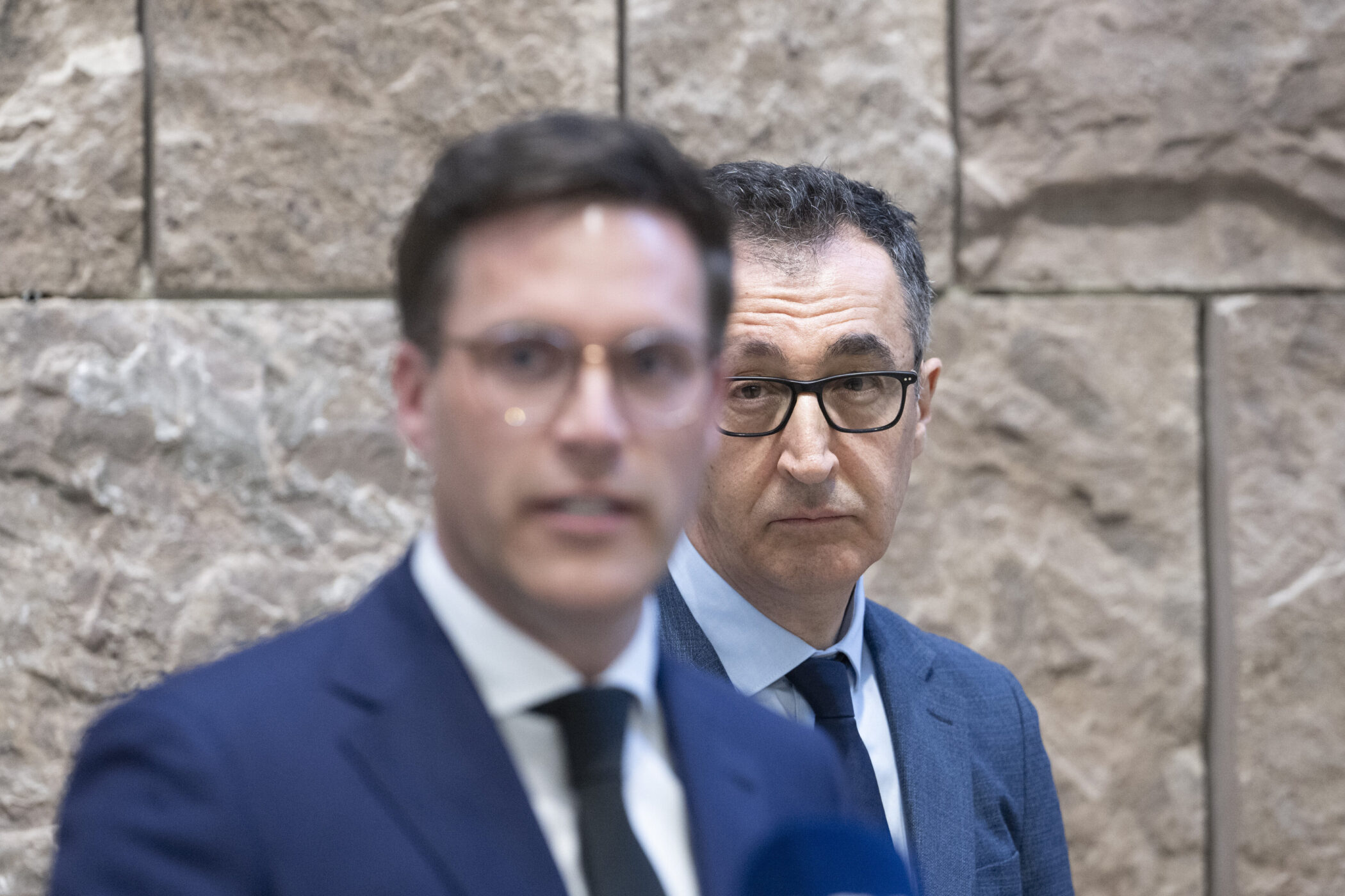 Zwei Männer in Anzügen vor einer Steinwand, einer unscharf im Vordergrund.