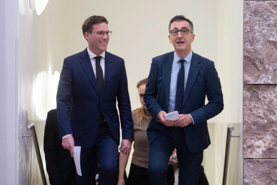 Zwei Männer in Anzügen gehen eine Treppe hinunter. Frau im Hintergrund.