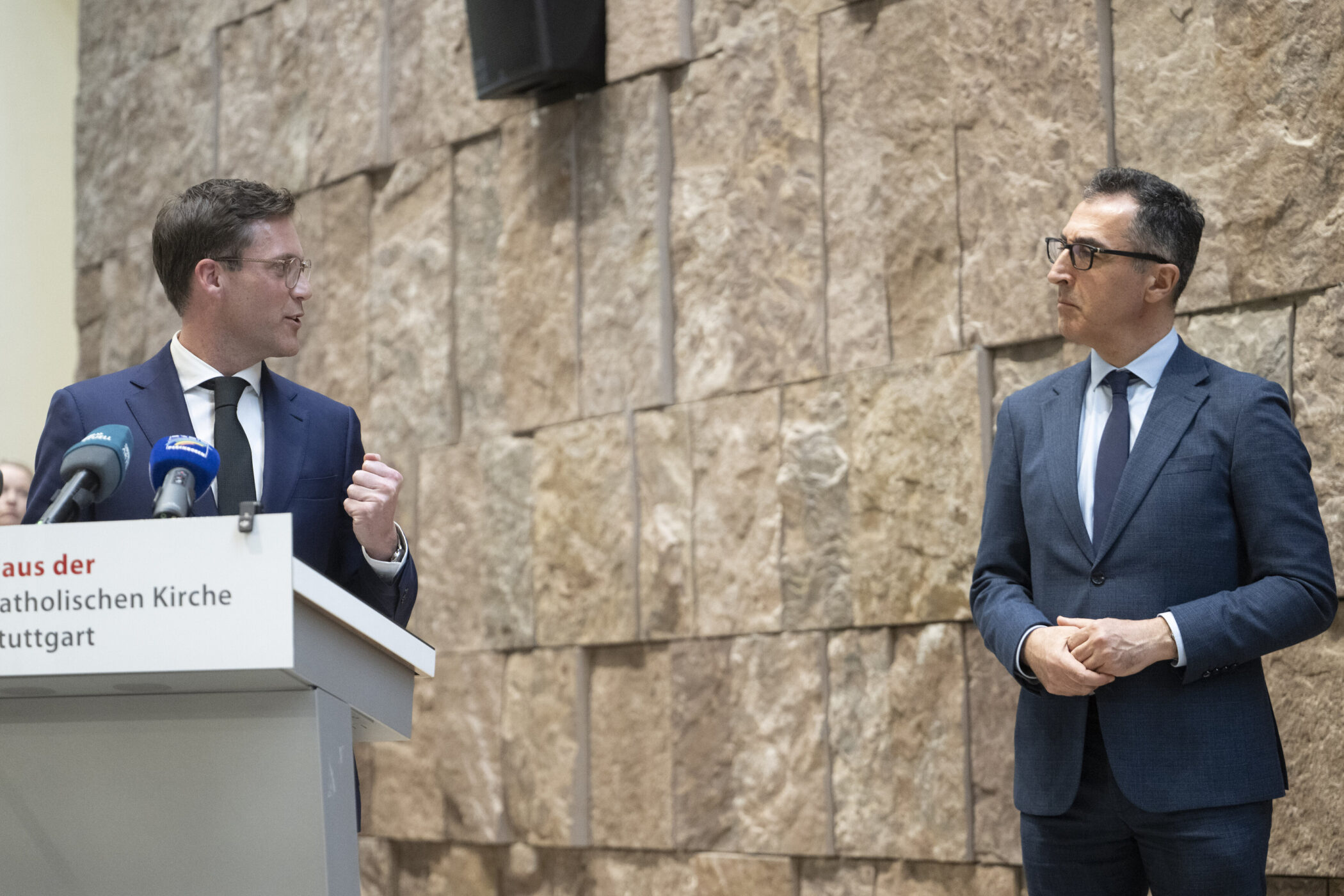 Zwei Männer in Anzügen stehen vor einer Steinwand, einer spricht am Pult mit Mikrofonen.
