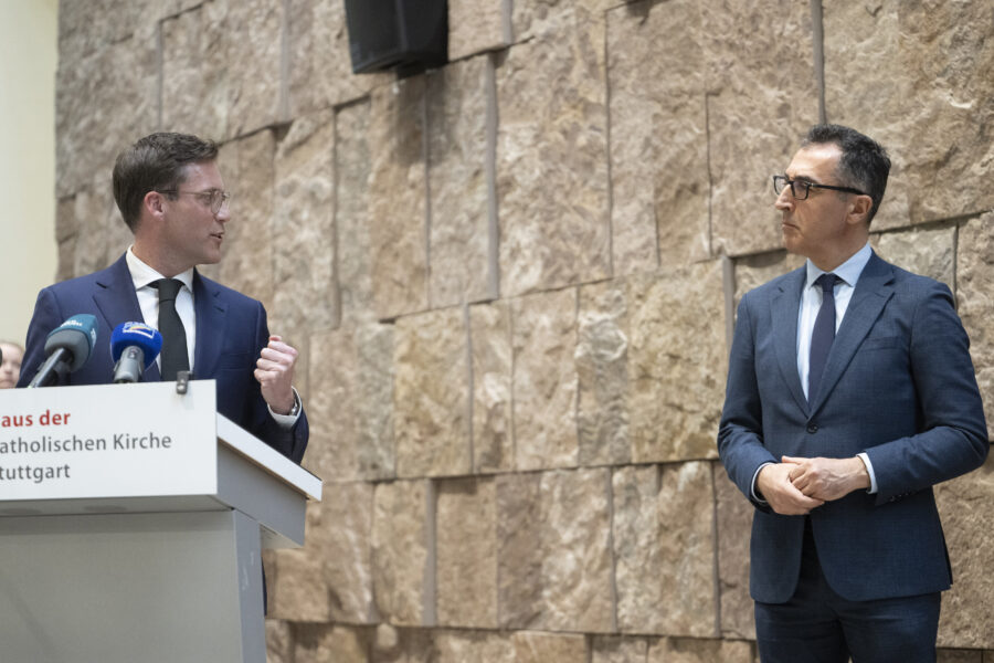 Zwei Männer in Anzügen stehen vor einer Steinwand, einer spricht am Pult mit Mikrofonen.