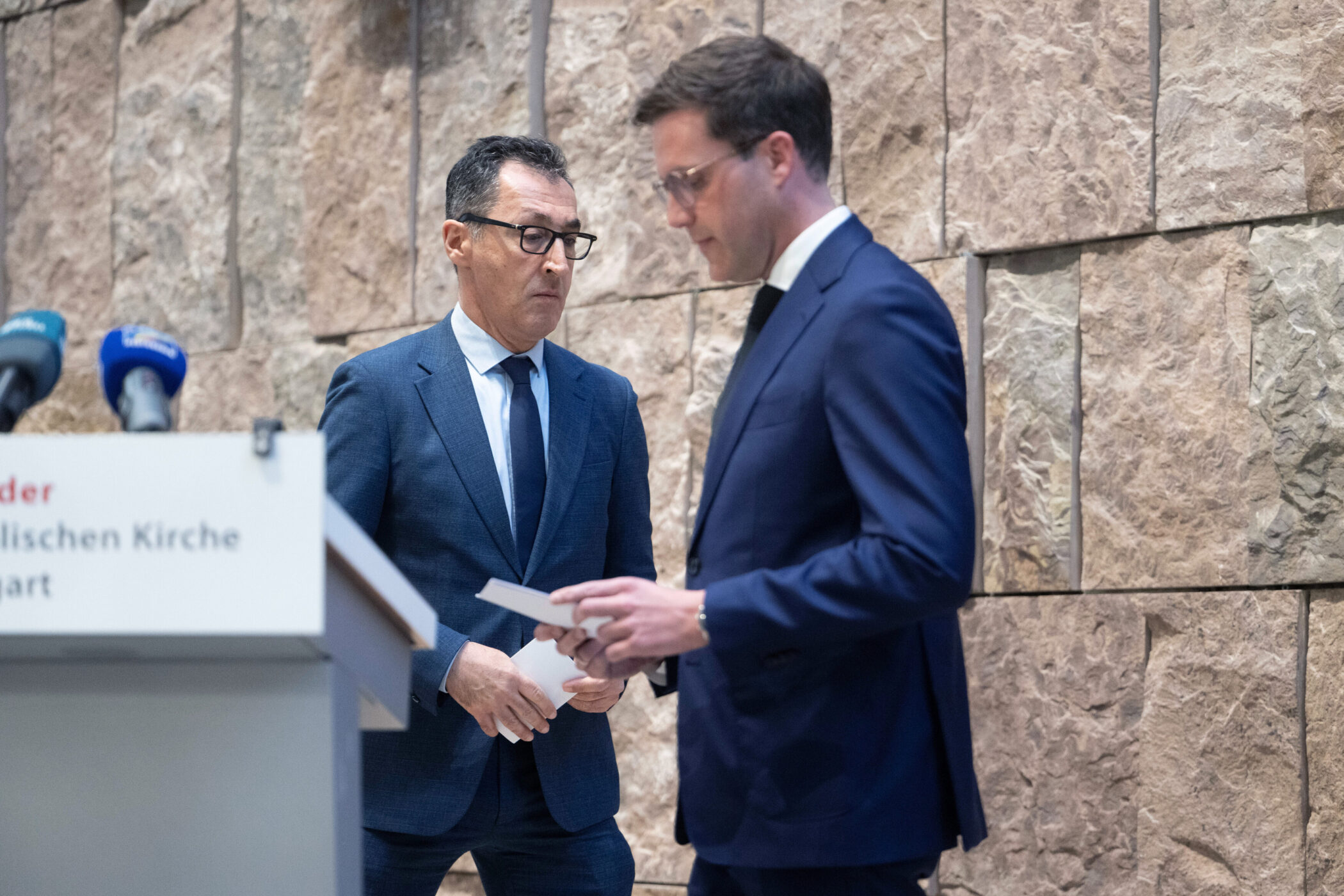 Zwei Männer in Anzügen stehen nebeneinander vor einer Steinwand.