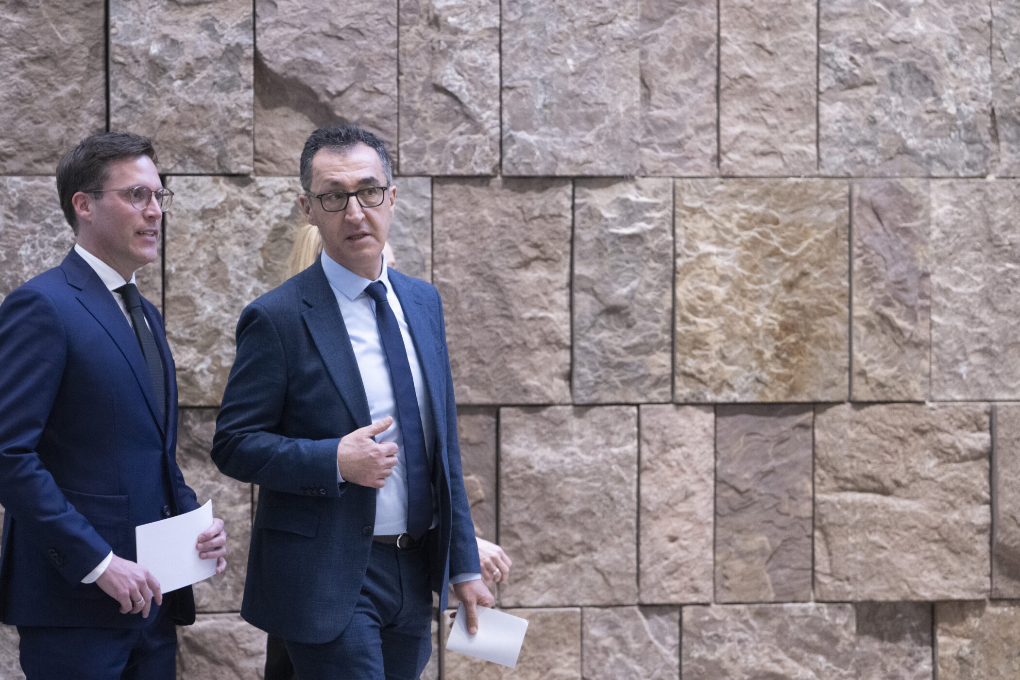 Zwei Männer in Anzügen mit Papier in der Hand vor Steinsakadenwand.