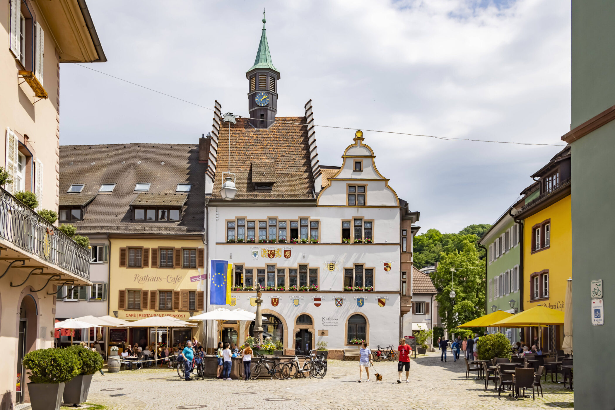 Pflasterstraße mit Menschen, Rathaus, Cafés und Fahrrädern auf einem Platz.