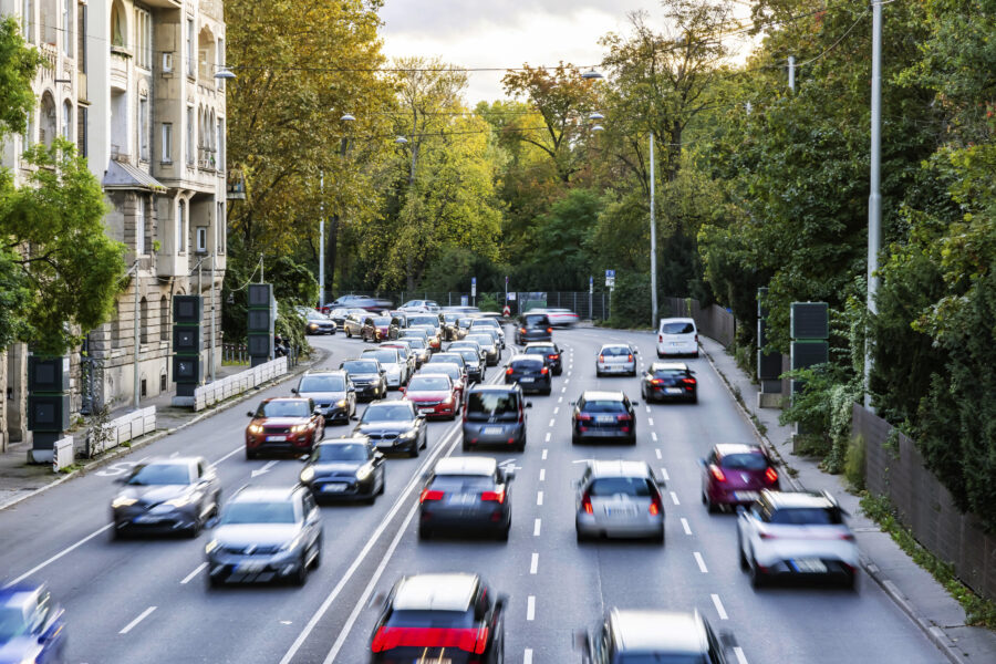 Straße mit dichtem Verkehr, Autos in beide Richtungen, Bäume im Hintergrund.