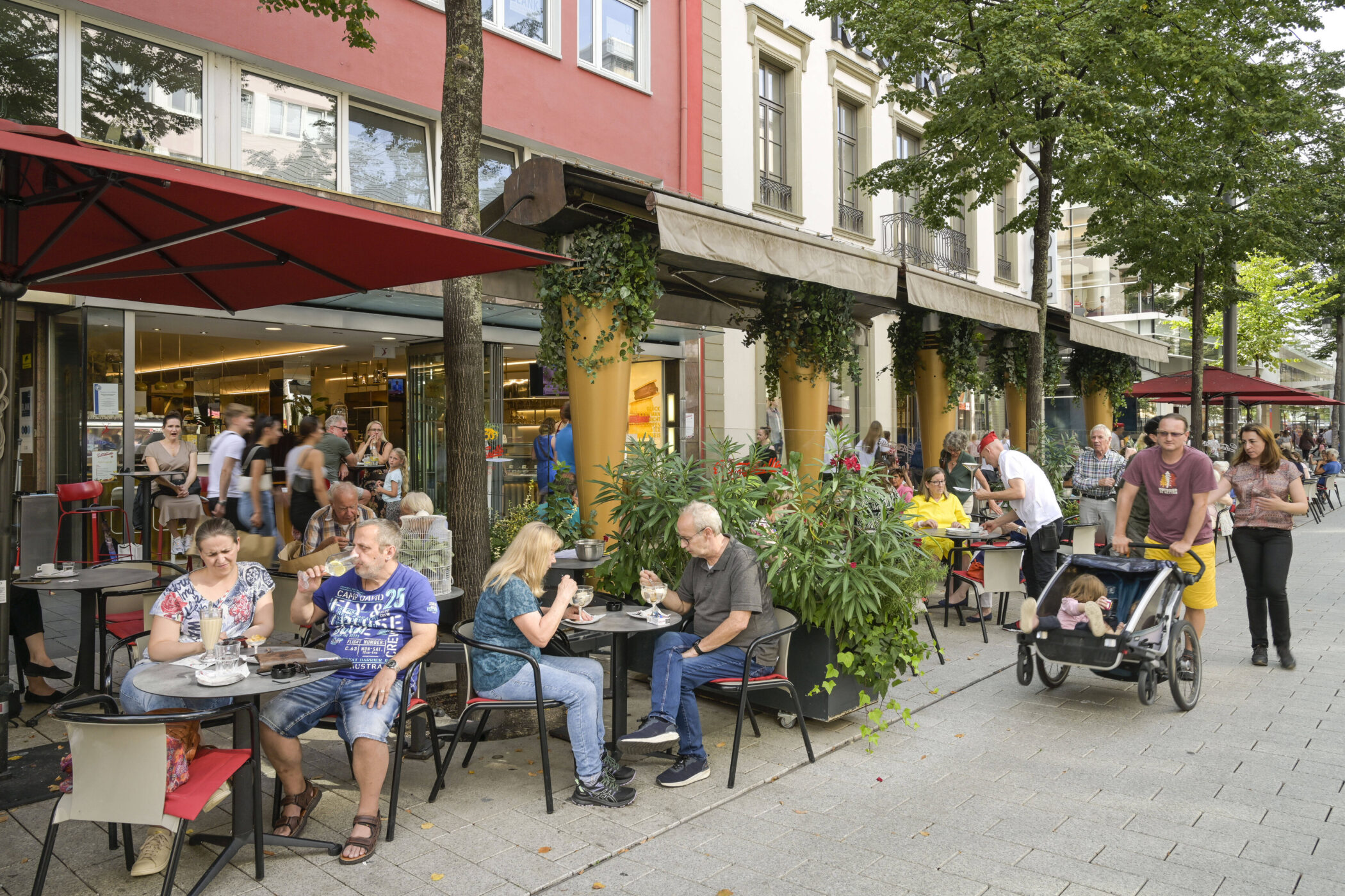 Mehrere Personen sitzen draußen an Café-Tischen unter roten Sonnenschirmen.
