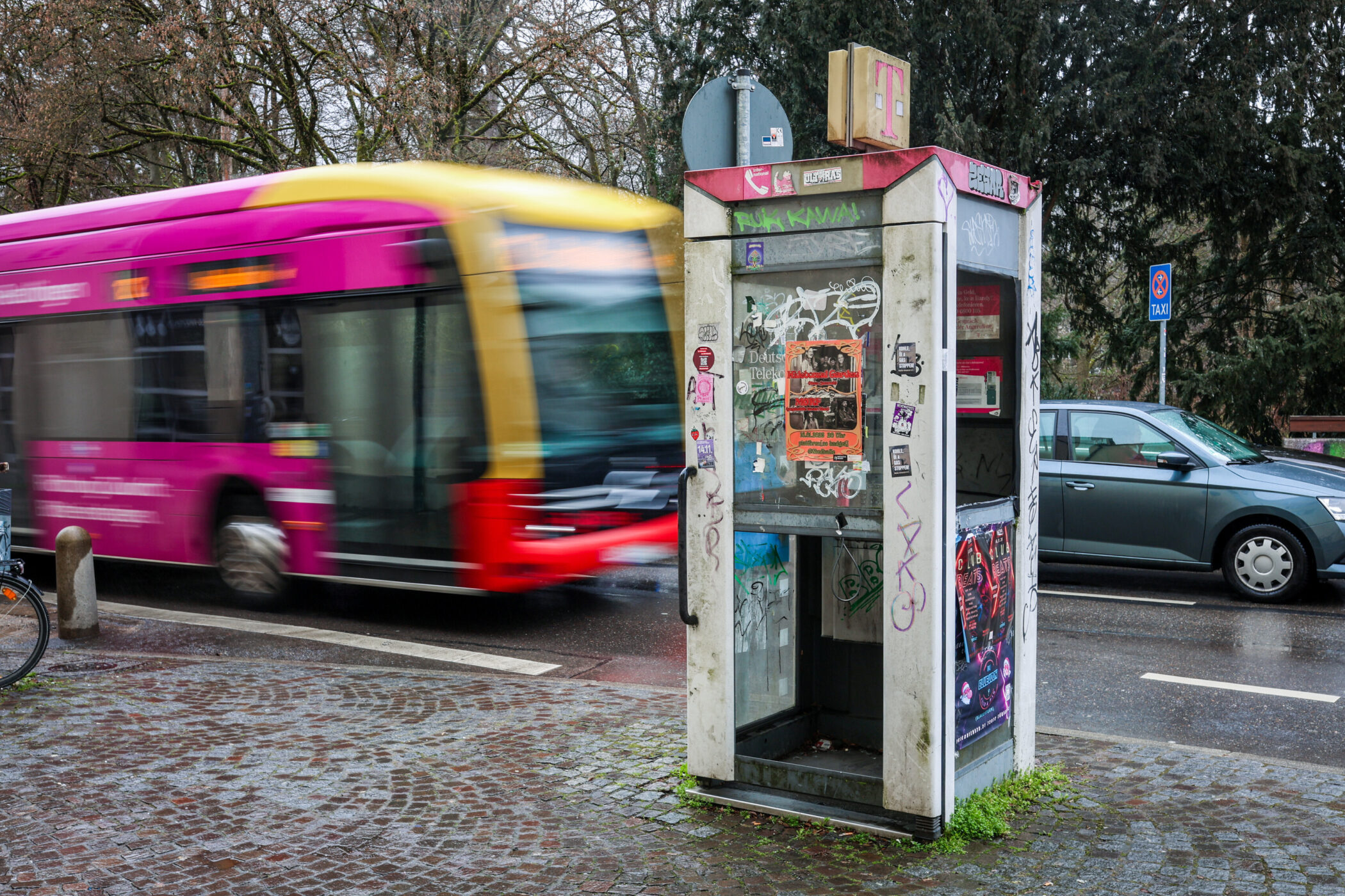 Alte Telefonzelle mit Graffiti und Stickern, vorbeifahrender Bus, parkendes Auto.
