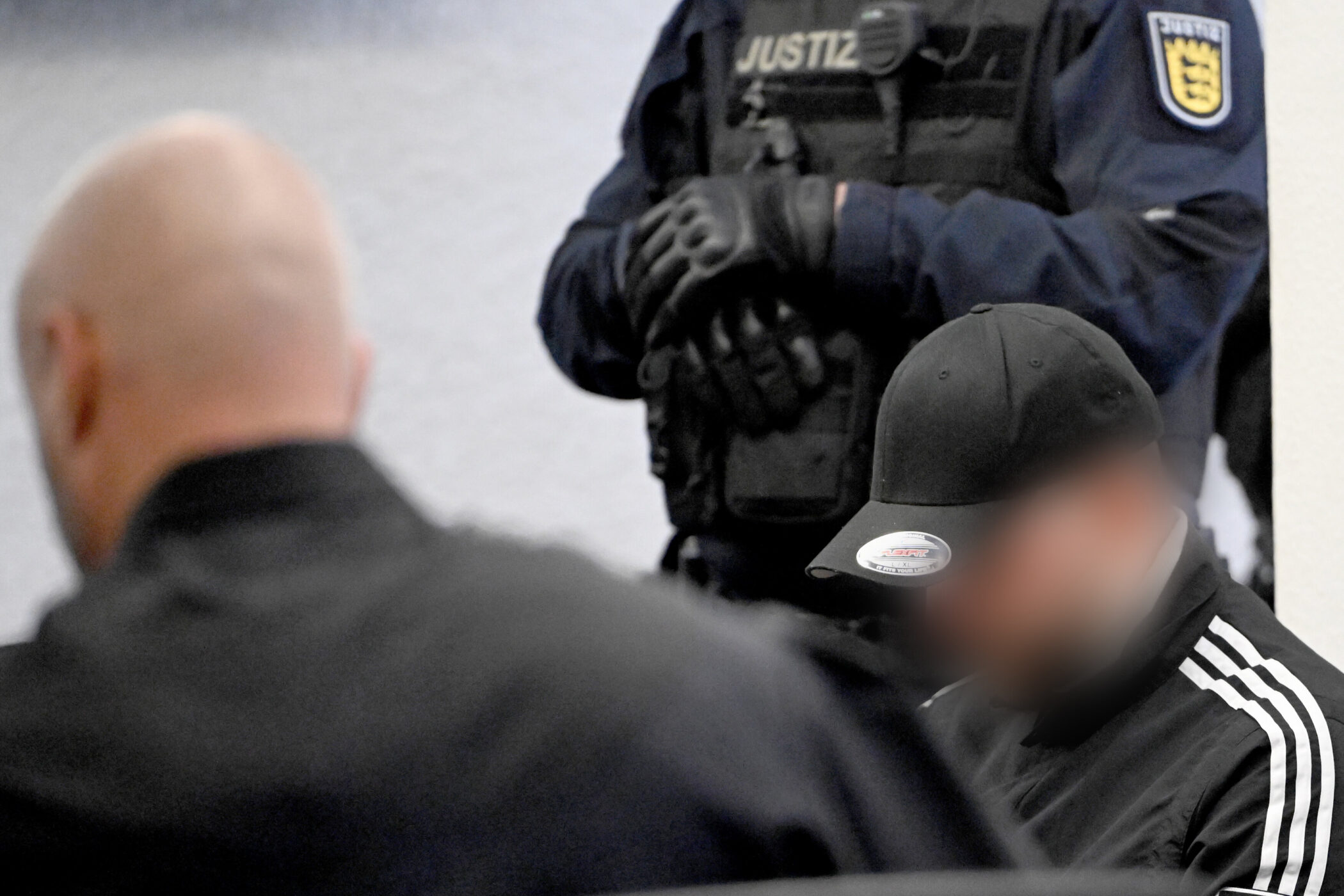 Drei Personen, unscharf im Vordergrund, Justizbeamter mit Weste im Hintergrund.