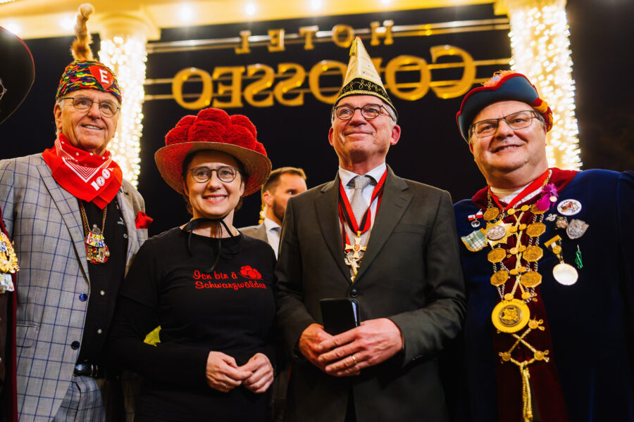 Vier Personen in festlicher Kleidung mit bunten Hüten und Medaillen, Schriftzug im Hintergrund.