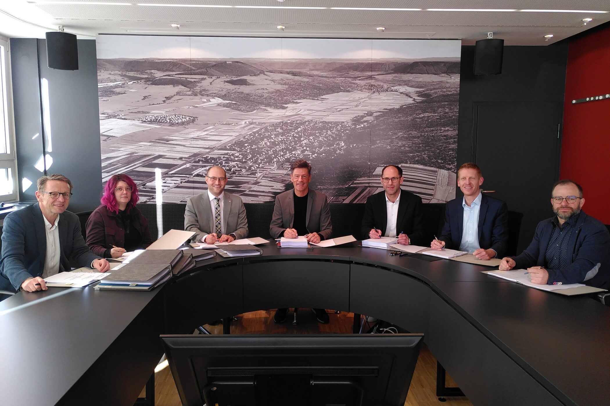 Acht Personen sitzen an einem gebogenen Tisch, hinter ihnen ein großes Landschaftsbild.