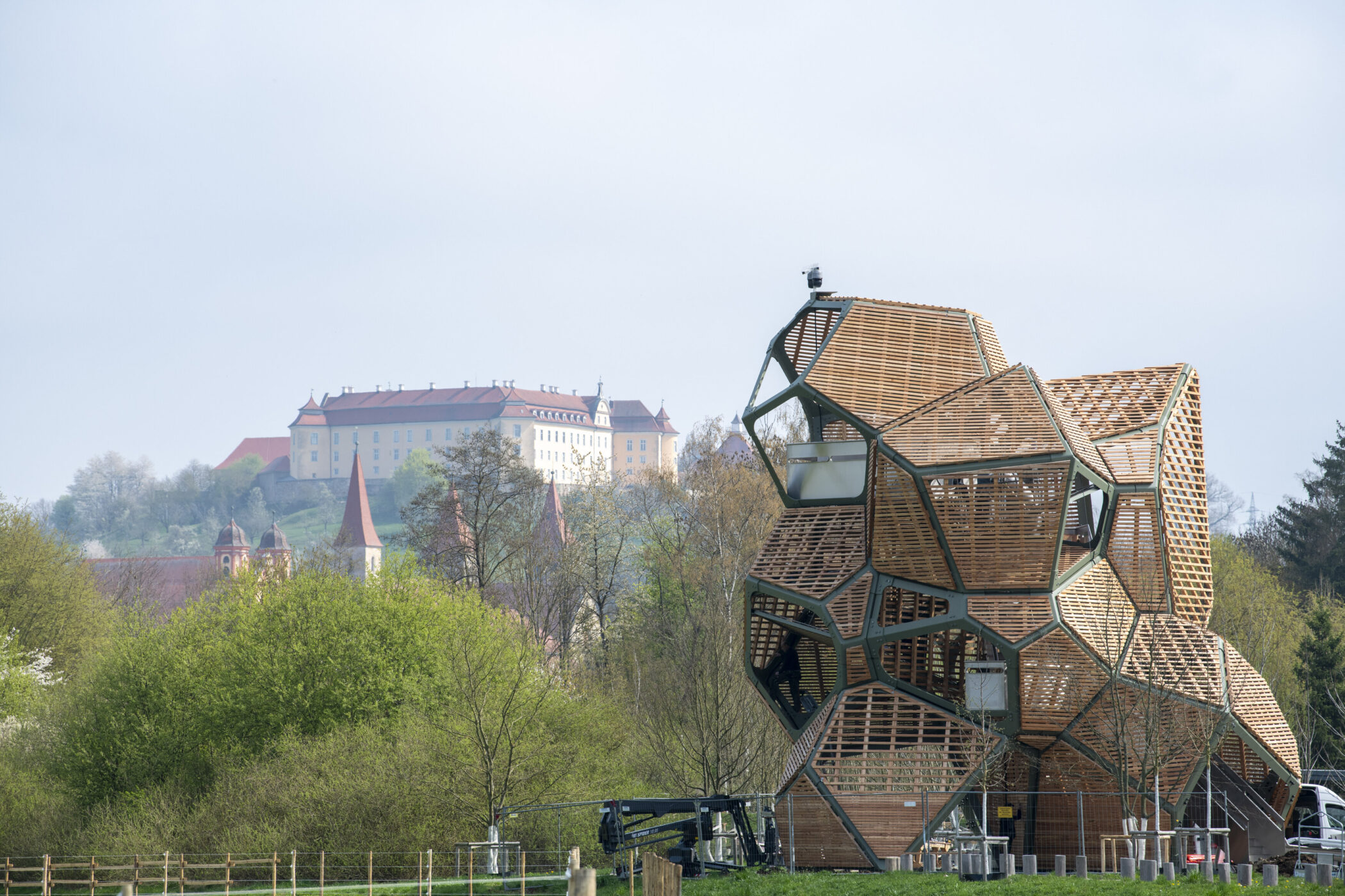 Holzstruktur im Vordergrund, Schloss im Hintergrund, umgeben von Bäumen und Wiesen.