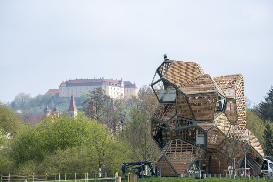Holzstruktur im Vordergrund, Schloss im Hintergrund, umgeben von Bäumen und Wiesen.