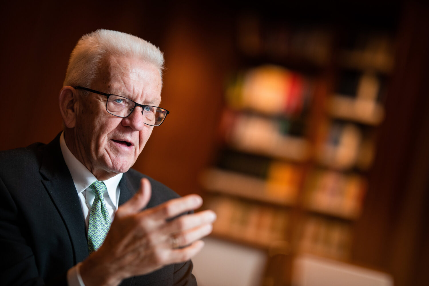 Älterer Mann mit Brille und Anzug gestikuliert, Hintergrund mit unscharfen Regalen.