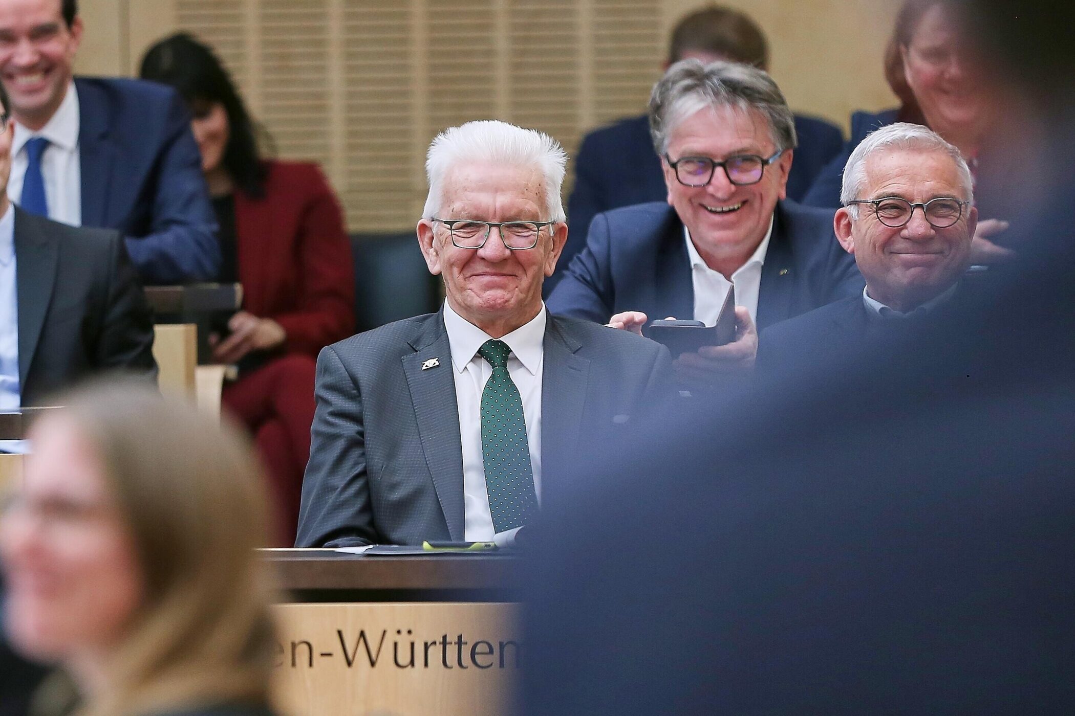 Drei Männer in Anzügen sitzen lächelnd in einem Raum.