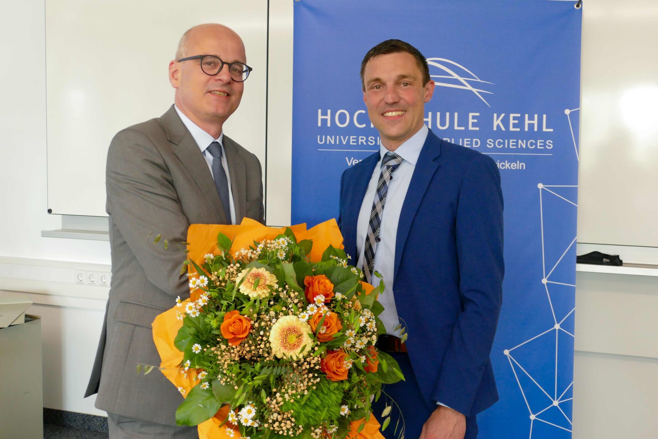 Zwei Männer im Anzug, einer hält Blumenstrauß, vor einem Hochschulbanner.