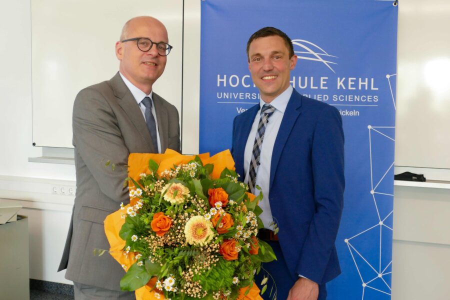 Zwei Männer im Anzug, einer hält Blumenstrauß, vor einem Hochschulbanner.