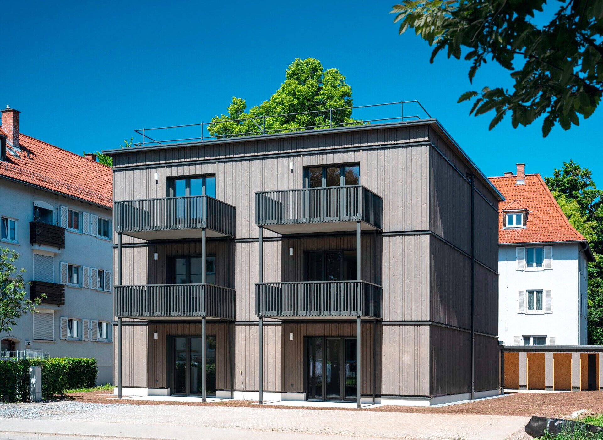 Modernes dreistöckiges Gebäude mit Balkonen, neben älteren Häusern mit roten Dächern.